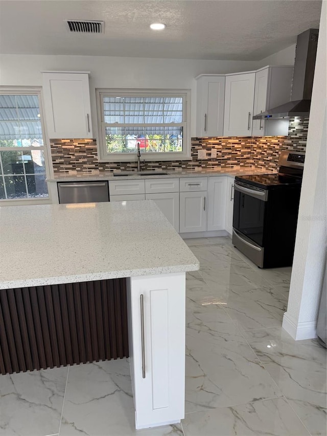 kitchen with light stone countertops, backsplash, stainless steel appliances, wall chimney range hood, and white cabinetry