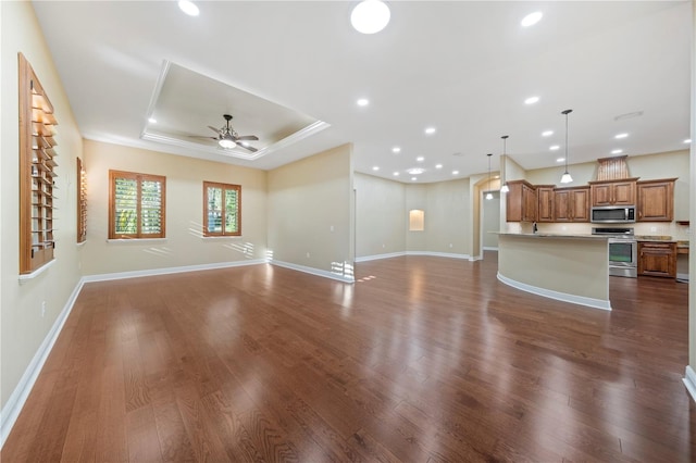 unfurnished living room with a tray ceiling, ceiling fan, crown molding, and dark wood-type flooring