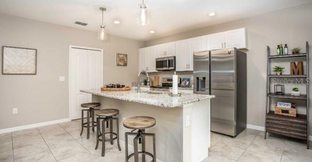 kitchen with light stone countertops, appliances with stainless steel finishes, decorative light fixtures, white cabinetry, and an island with sink