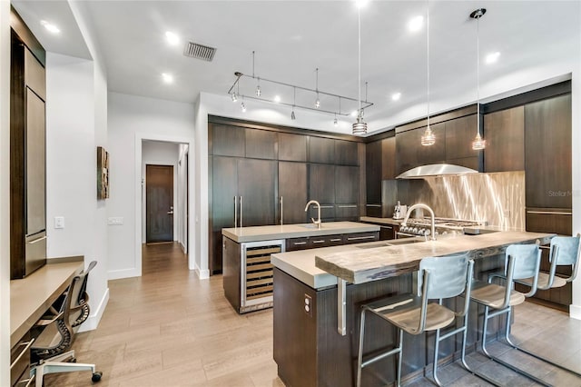 kitchen featuring beverage cooler, backsplash, an island with sink, decorative light fixtures, and dark brown cabinets