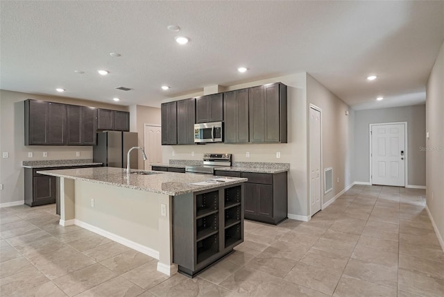 kitchen featuring appliances with stainless steel finishes, light stone counters, dark brown cabinets, sink, and an island with sink