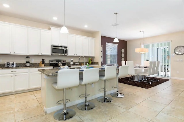 kitchen featuring stainless steel appliances, dark stone countertops, decorative light fixtures, a center island with sink, and white cabinets