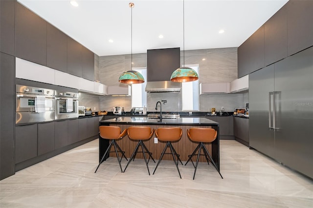 kitchen with tasteful backsplash, wall chimney exhaust hood, stainless steel appliances, a kitchen island with sink, and hanging light fixtures