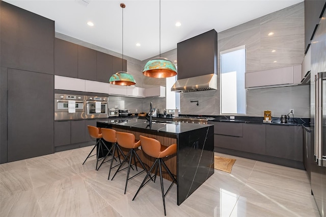 kitchen with a kitchen bar, appliances with stainless steel finishes, a kitchen island with sink, wall chimney range hood, and pendant lighting