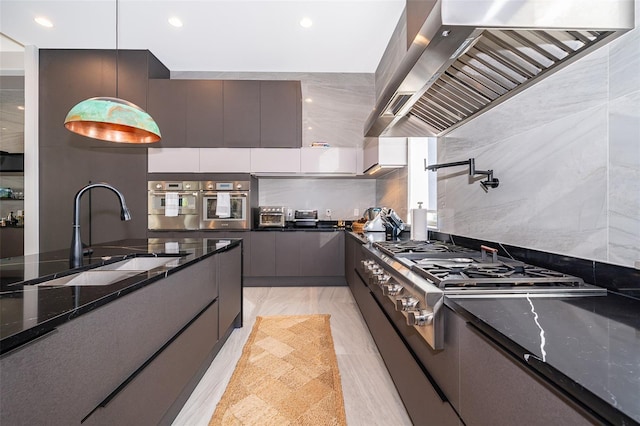 kitchen featuring stainless steel oven, sink, wall chimney range hood, dark stone counters, and decorative light fixtures