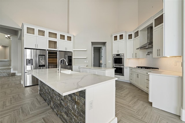 kitchen with appliances with stainless steel finishes, wall chimney range hood, a center island with sink, white cabinets, and a high ceiling