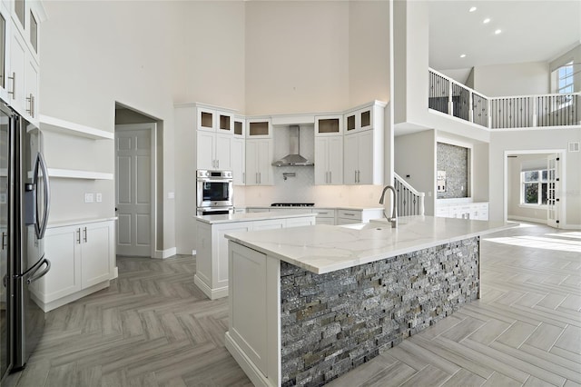 kitchen with a towering ceiling, stainless steel appliances, wall chimney range hood, white cabinets, and an island with sink