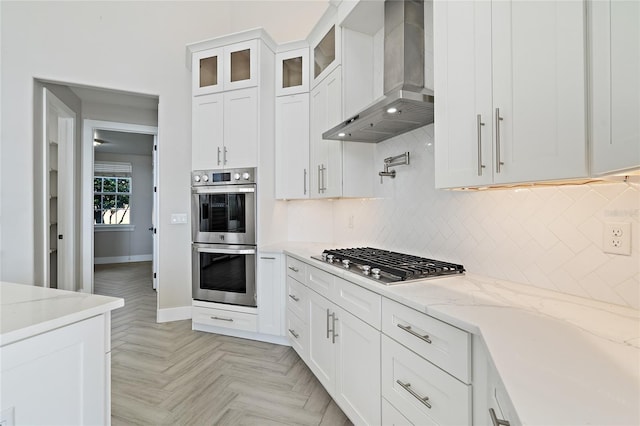 kitchen with wall chimney exhaust hood, light stone counters, decorative backsplash, white cabinets, and appliances with stainless steel finishes