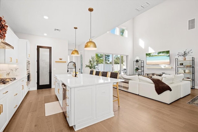 kitchen featuring light wood-type flooring, stainless steel appliances, decorative light fixtures, a center island with sink, and white cabinetry