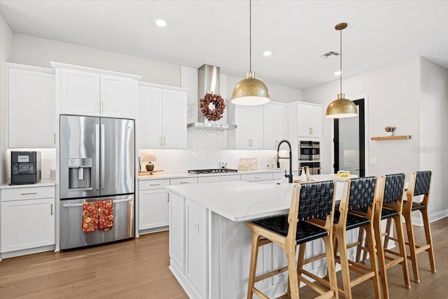 kitchen with white cabinetry, wall chimney exhaust hood, stainless steel appliances, decorative light fixtures, and a center island with sink