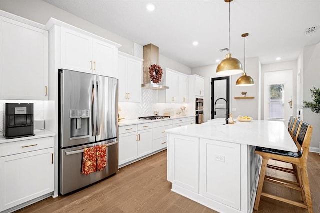 kitchen featuring wall chimney exhaust hood, white cabinets, an island with sink, and stainless steel appliances