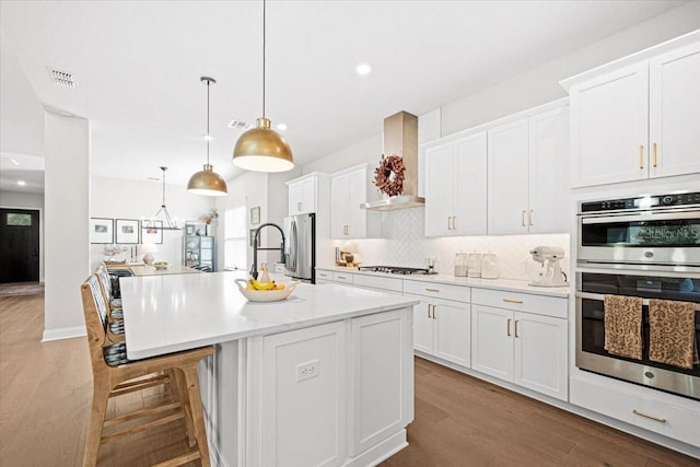 kitchen with pendant lighting, a center island with sink, wall chimney range hood, white cabinetry, and stainless steel appliances