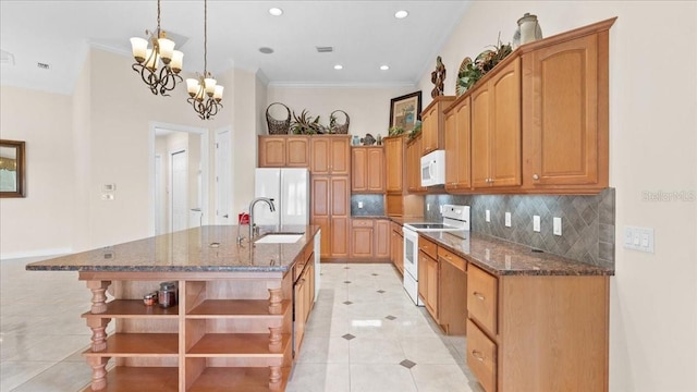 kitchen with sink, an inviting chandelier, dark stone countertops, an island with sink, and white appliances