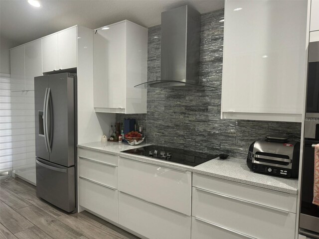 kitchen with stainless steel refrigerator with ice dispenser, wall chimney exhaust hood, black electric cooktop, light hardwood / wood-style floors, and white cabinetry