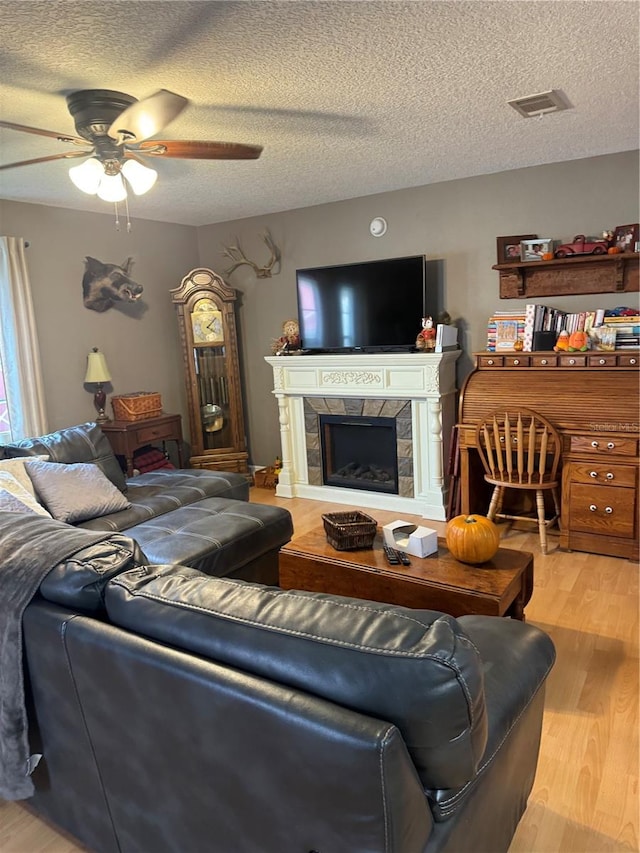 living room featuring ceiling fan, a stone fireplace, light wood-type flooring, and a textured ceiling