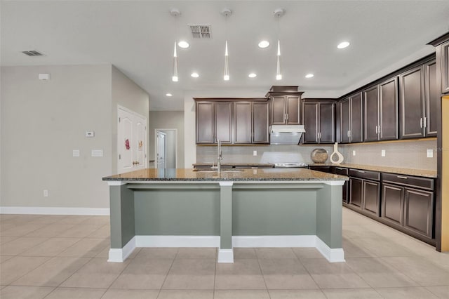 kitchen with light tile patterned floors, an island with sink, stone counters, dark brown cabinetry, and sink