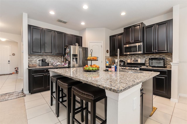 kitchen with appliances with stainless steel finishes, a center island with sink, light tile patterned floors, and tasteful backsplash