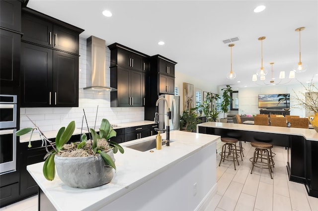 kitchen featuring decorative backsplash, wall chimney range hood, a large island with sink, hanging light fixtures, and a breakfast bar area