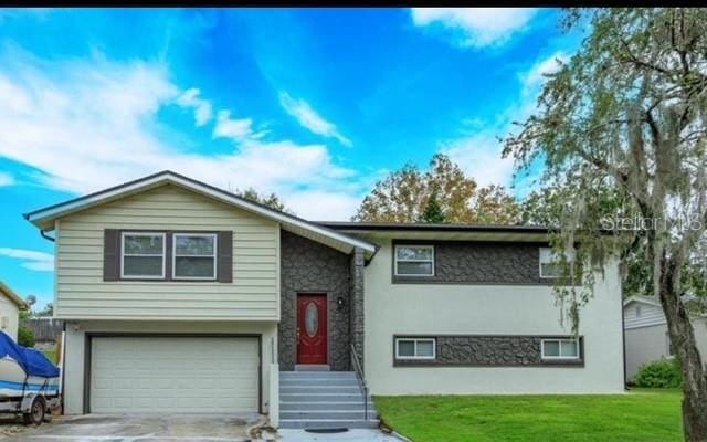 bi-level home featuring stucco siding, an attached garage, stone siding, driveway, and a front lawn