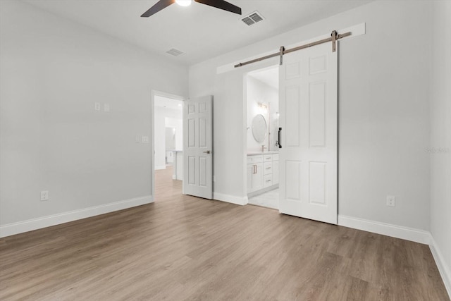 unfurnished bedroom with ensuite bathroom, a barn door, light wood-type flooring, and ceiling fan