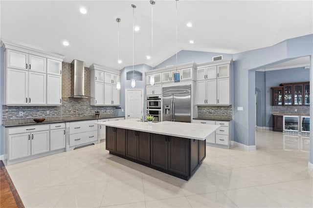 kitchen featuring stainless steel appliances, wall chimney range hood, vaulted ceiling, decorative light fixtures, and a center island with sink