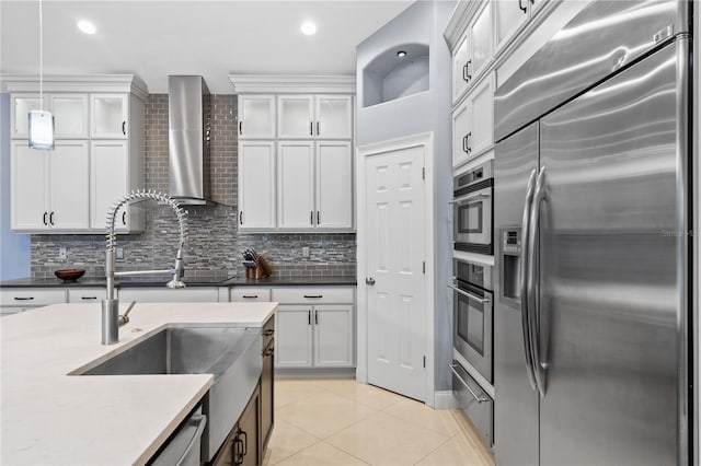 kitchen featuring stainless steel appliances, wall chimney range hood, light tile patterned floors, decorative light fixtures, and white cabinetry