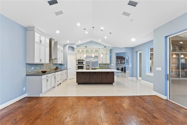 kitchen featuring appliances with stainless steel finishes, tasteful backsplash, wall chimney range hood, hanging light fixtures, and an island with sink