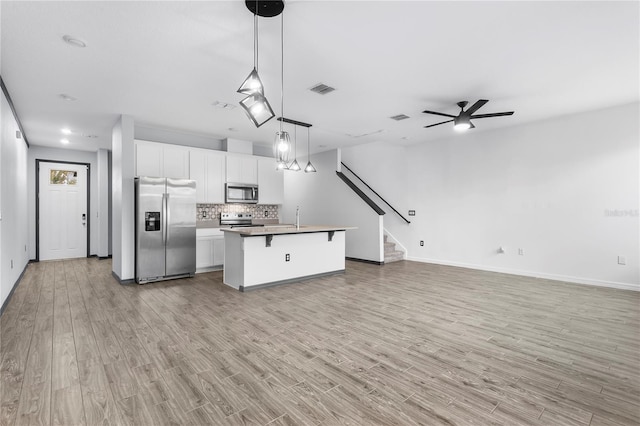 kitchen with hanging light fixtures, appliances with stainless steel finishes, a kitchen island with sink, white cabinets, and light wood-type flooring