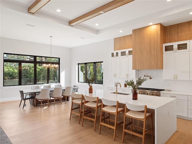 kitchen featuring white cabinets, beamed ceiling, hanging light fixtures, a kitchen island with sink, and a notable chandelier