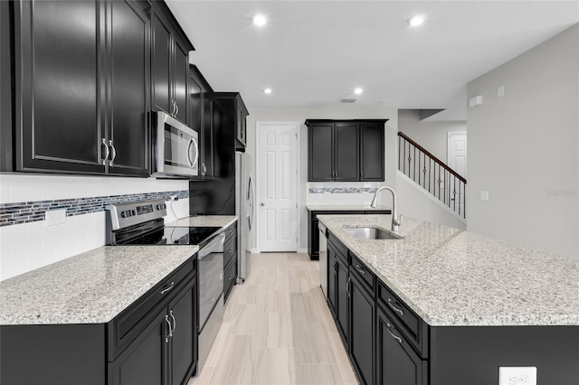 kitchen with a kitchen island with sink, sink, tasteful backsplash, light stone counters, and stainless steel appliances