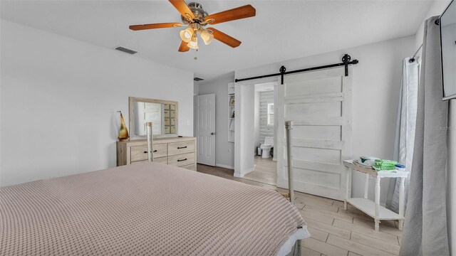 bedroom with light wood-type flooring, a barn door, ceiling fan, and connected bathroom