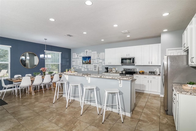 kitchen with white cabinets, a center island with sink, hanging light fixtures, light stone countertops, and appliances with stainless steel finishes