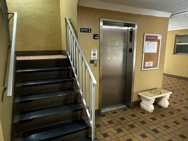 stairway with brick floor, elevator, and a textured ceiling