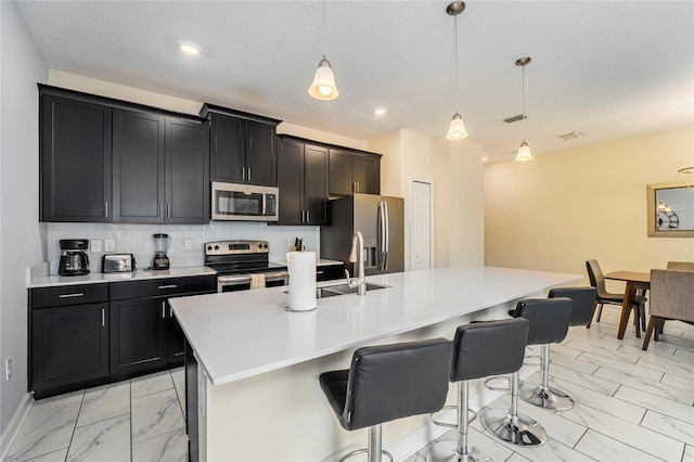 kitchen featuring tasteful backsplash, decorative light fixtures, a kitchen bar, a center island with sink, and appliances with stainless steel finishes