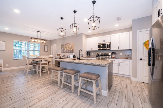 kitchen featuring a center island with sink, hanging light fixtures, light stone counters, white cabinetry, and stainless steel appliances