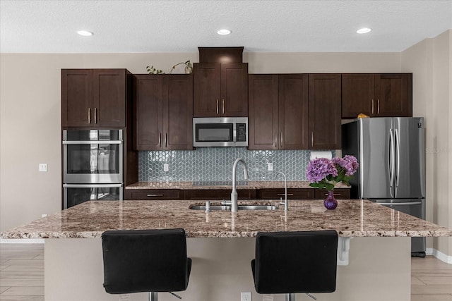 kitchen featuring a breakfast bar, a kitchen island with sink, light stone countertops, a textured ceiling, and appliances with stainless steel finishes