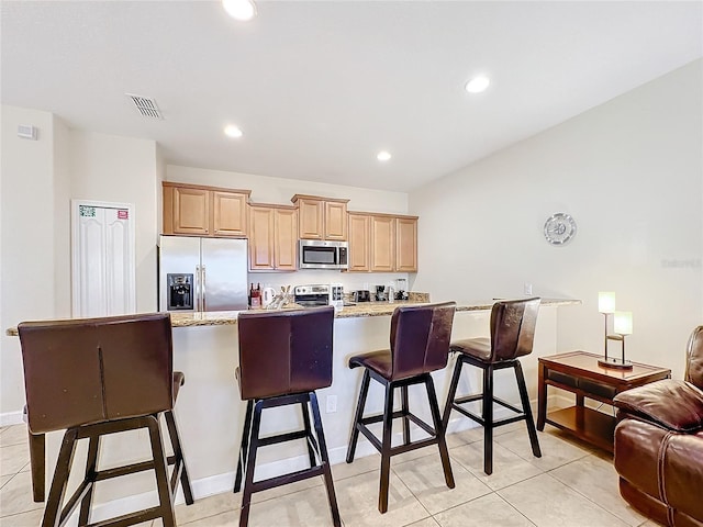 kitchen featuring light stone countertops, a breakfast bar area, light brown cabinetry, light tile patterned floors, and appliances with stainless steel finishes