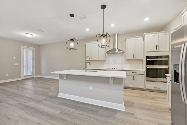 kitchen with wall chimney exhaust hood, sink, a center island with sink, white cabinets, and hanging light fixtures