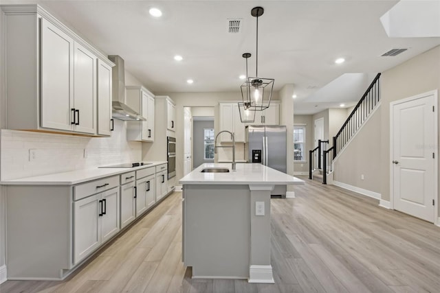 kitchen featuring sink, wall chimney range hood, a center island with sink, light hardwood / wood-style flooring, and hanging light fixtures