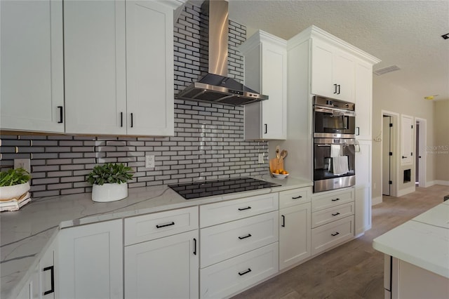 kitchen featuring white cabinets, light stone counters, black electric stovetop, wall chimney range hood, and double oven