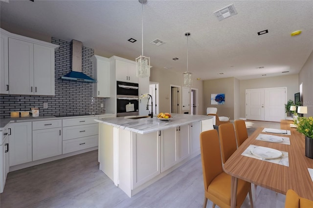 kitchen with wall chimney exhaust hood, hanging light fixtures, an island with sink, and visible vents