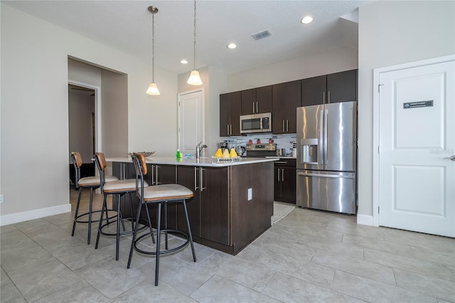 kitchen with a center island with sink, a kitchen breakfast bar, hanging light fixtures, appliances with stainless steel finishes, and dark brown cabinets