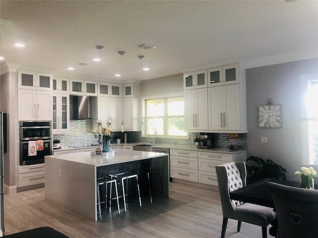 kitchen with wall chimney exhaust hood, double oven, white cabinets, a kitchen island, and hanging light fixtures