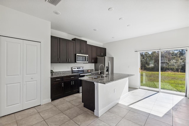 kitchen with dark brown cabinetry, dark stone counters, a kitchen island with sink, light tile patterned flooring, and appliances with stainless steel finishes