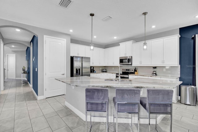 kitchen with hanging light fixtures, stainless steel appliances, light stone counters, a kitchen island with sink, and white cabinets