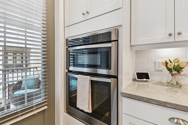 kitchen with double oven, backsplash, white cabinetry, and light stone countertops