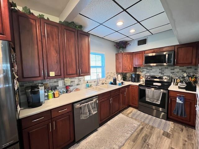 kitchen with a drop ceiling, sink, light hardwood / wood-style flooring, decorative backsplash, and stainless steel appliances