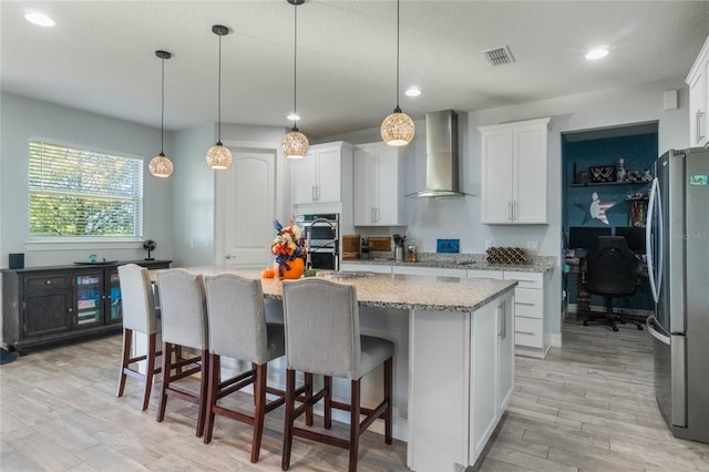 kitchen featuring a kitchen island with sink, white cabinets, wall chimney range hood, and appliances with stainless steel finishes