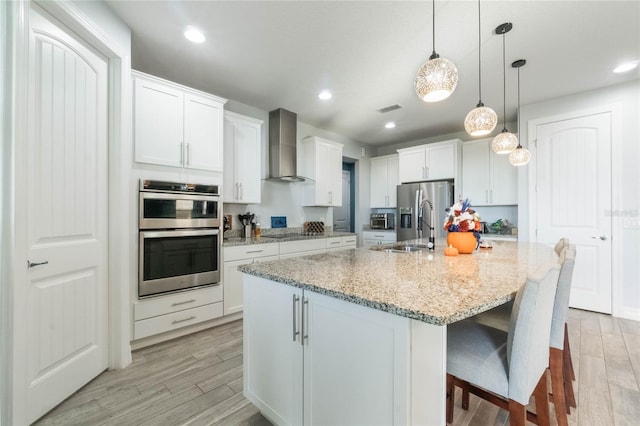 kitchen featuring white cabinets, wall chimney exhaust hood, light wood-type flooring, an island with sink, and stainless steel appliances