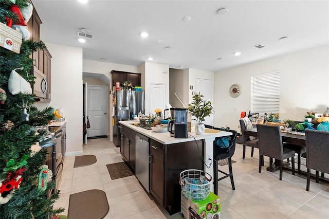 kitchen featuring a breakfast bar, light tile patterned floors, an island with sink, dark brown cabinets, and stainless steel appliances